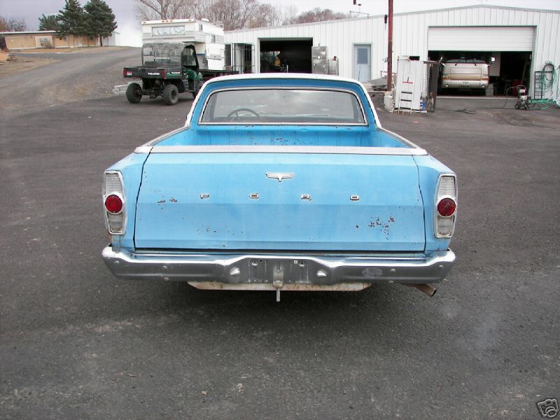 Rear view of light blue Ford Ranchero with FORD tailgate lettering, parked on gravel near white shop buildings