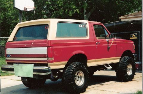 1990 Ford Bronco - This lifted Ford Bronco, painted red with tan accents, is parked outdoors near a garage and basketball hoop.