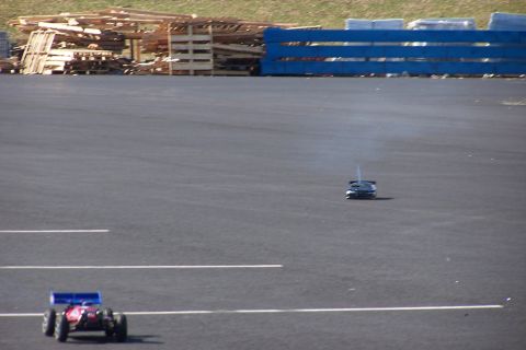 1994 Ford Bronco - Two remote control cars are seen racing on an asphalt track, with a background of stacked pallets and blue fencing.
