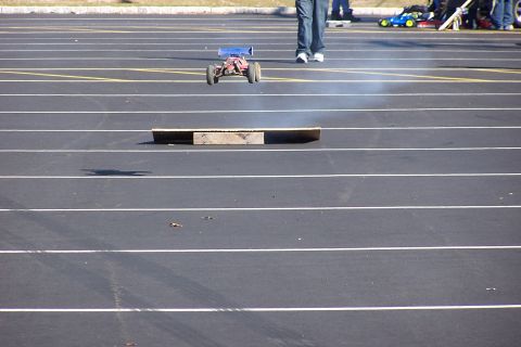 1994 Ford Bronco - A remote control car performs a jump over a wooden obstacle on a paved track, with spectators in the background.