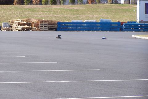 1994 Ford Bronco - Two remote control cars are visible on an asphalt surface, with a background of stacked pallets and blue barriers.