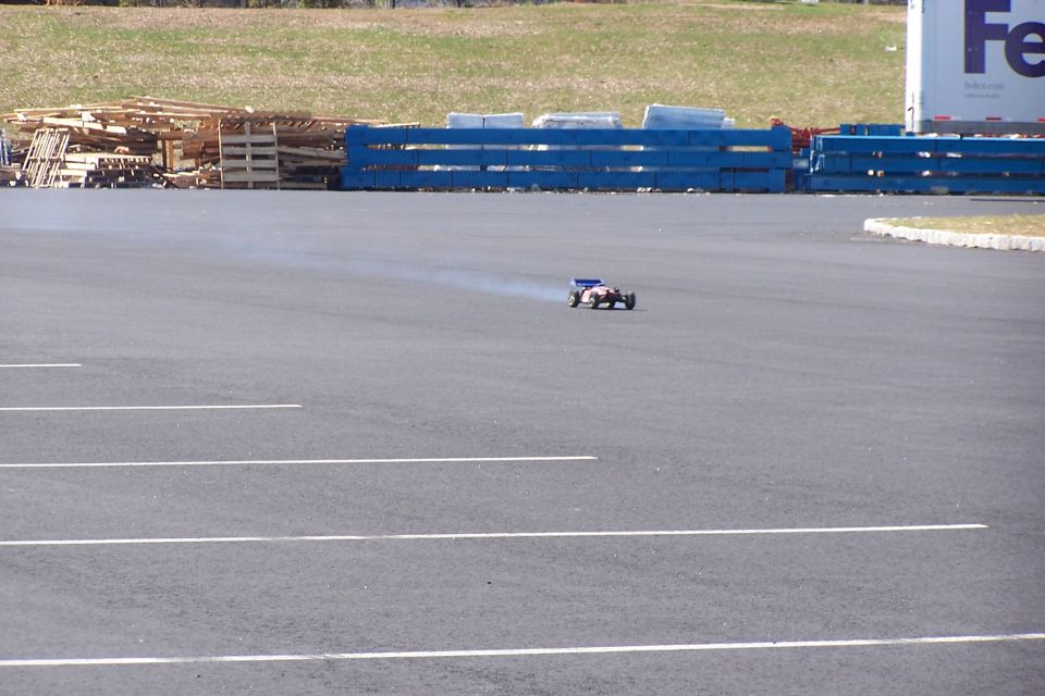 Remote-controlled car on asphalt with tire smoke, blue barrier, and FedEx truck in background