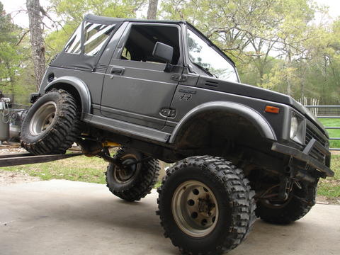 1987 Ford Bronco - This gray off-road SUV is shown with its front end lifted on a ramp, highlighting its modified suspension and large tires.
