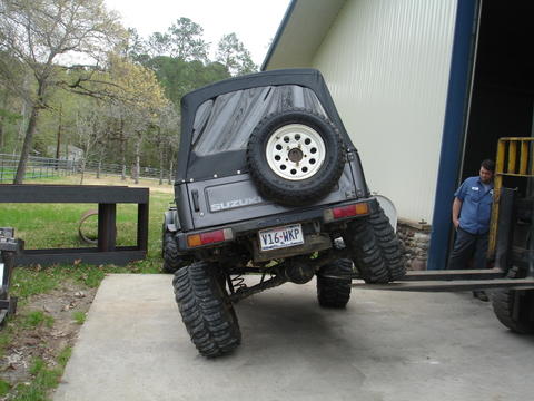 1987 Ford Bronco - A Suzuki Samurai is being lifted by a forklift, showing its rear end and large off-road tires.
