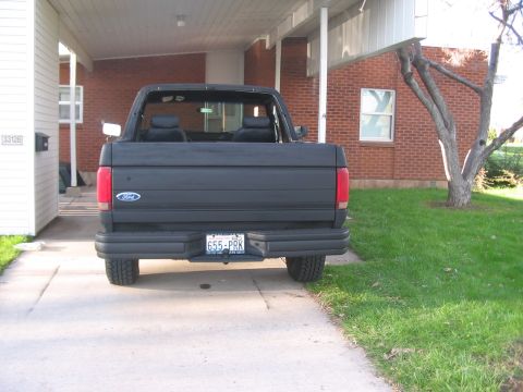 1991 Ford Bronco - The rear of a black Ford Bronco is visible, parked under a carport next to a brick house.
