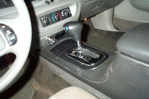 1999 Ford Crown Victoria - Interior center console of a gray-trimmed vehicle showing a black automatic floor shifter with chrome shaft, an aftermarket single-DIN ster…