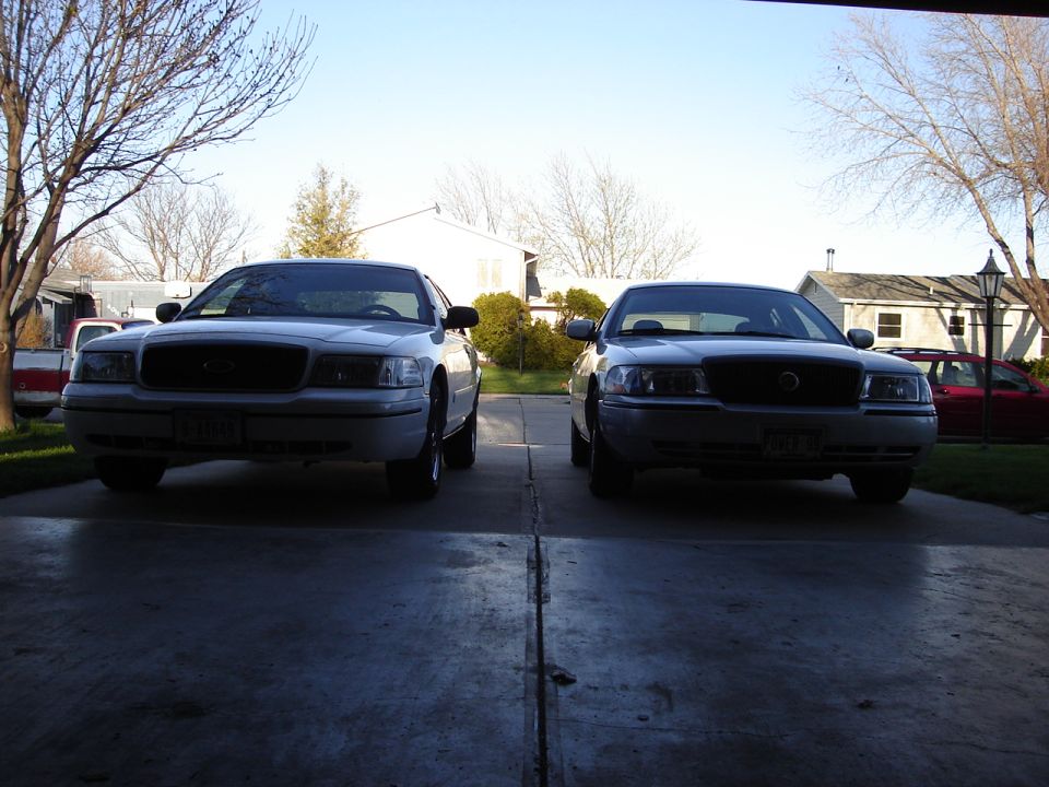 Two Ford Crown Victorias parked in a driveway, one white and one gray. The white one is on the left, gray on the right