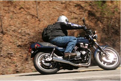 1985 Ford LTD Crown Victoria - Rider on a motorcycle navigates a curve on a road bordered by a wooded slope.
