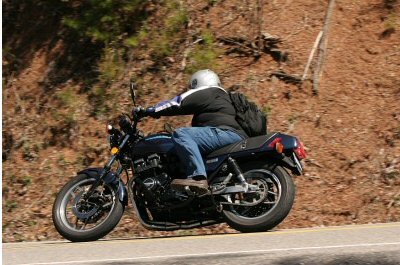 1985 Ford LTD Crown Victoria - A rider on a dark blue motorcycle navigates a scenic road, with a wooded hillside in the background.