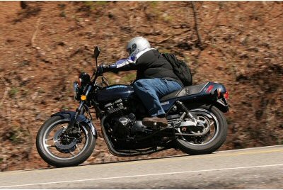 1985 Ford LTD Crown Victoria - Rider on a motorcycle navigates a curve on a road bordered by a dirt embankment.