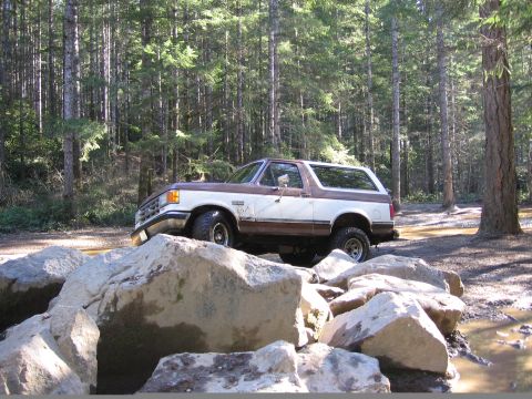 1987 Ford Bronco - A two-tone brown and white 1987 Ford Bronco is shown off-roading over large rocks in a wooded area.
