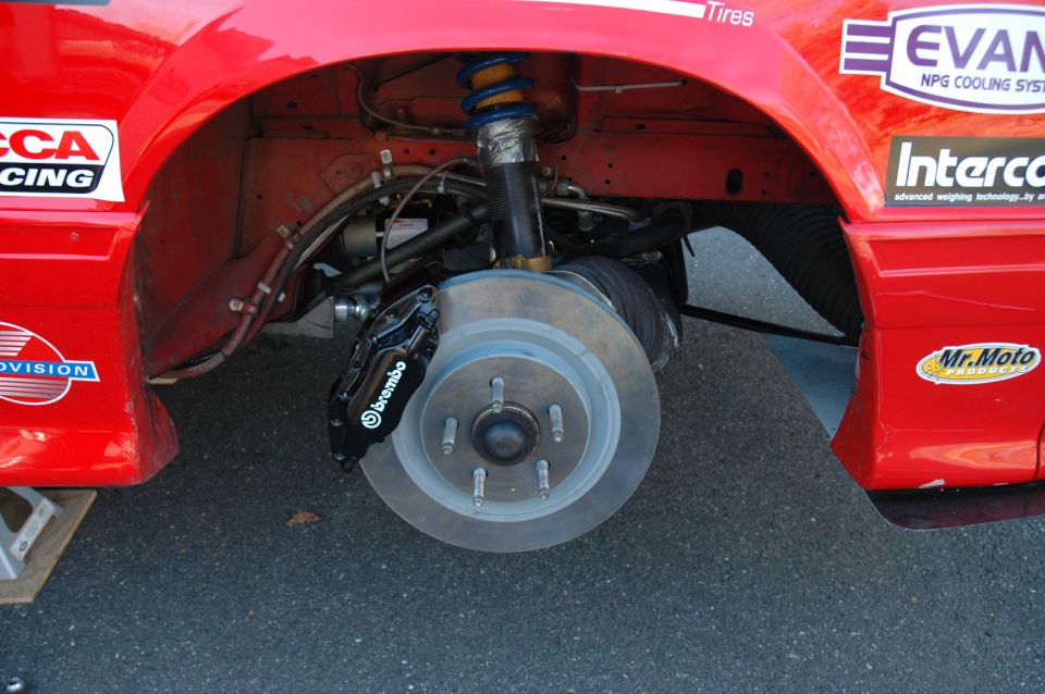 Close-up of a red race car's front wheel, showing a Brembo brake caliper and rotor. Racing decals are visible on the fender