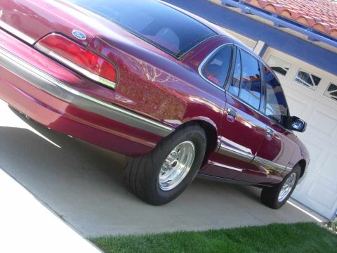 1992 Ford Crown Victoria - Low, tilted 3/4 rear shot of a deep maroon Ford Crown Victoria sedan on a concrete driveway, showing the full-width taillight panel, Ford o…