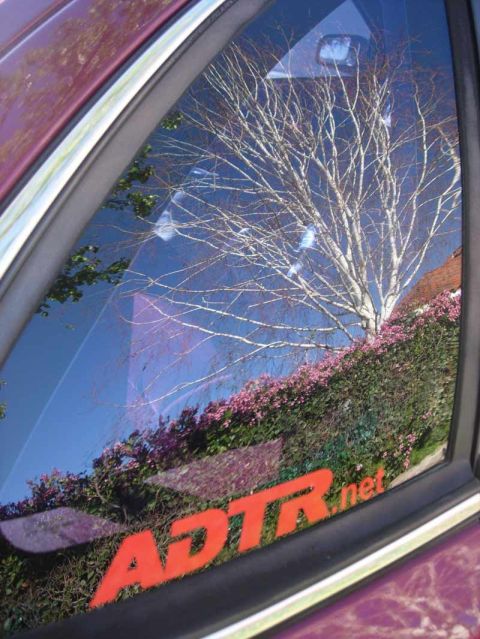 1992 Ford Crown Victoria - Close-up of a maroon car's rear quarter window carrying an orange 'ADTR.net' decal along the bottom of the glass, with a bare tree, blue sk…