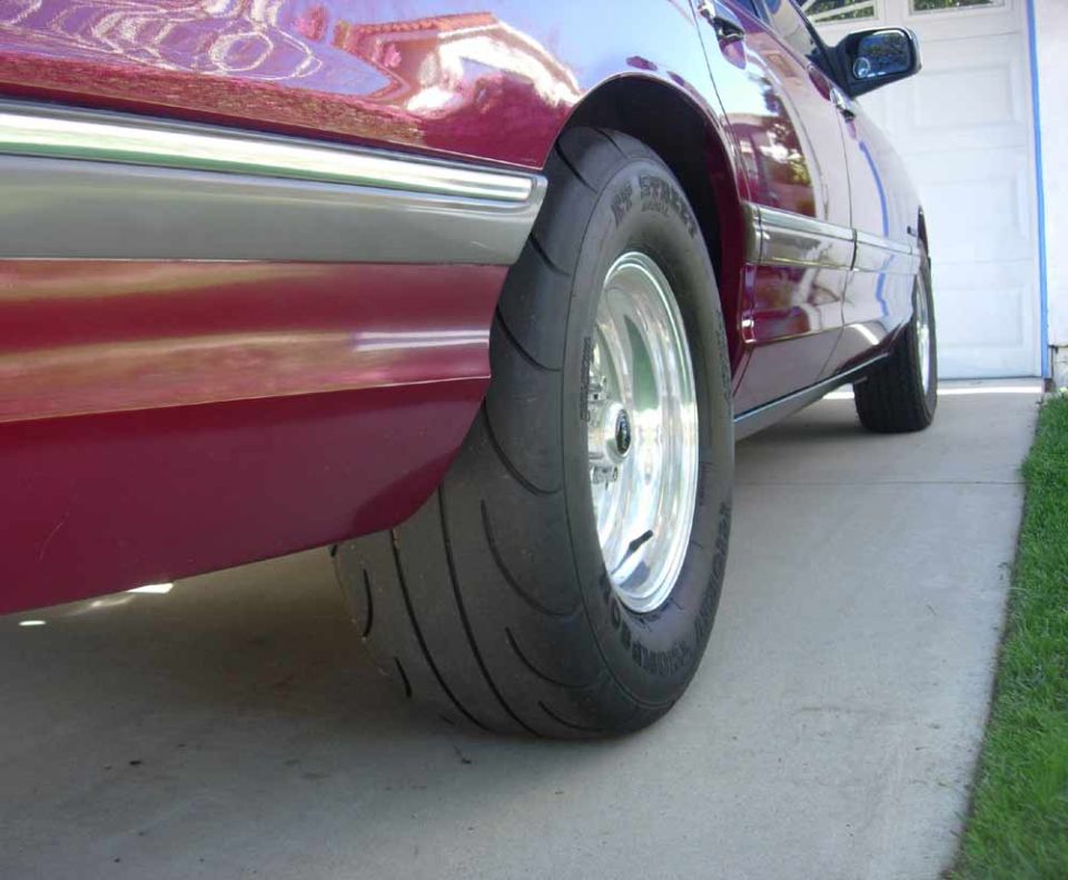 Maroon sedan rear wheel with Mickey Thompson drag radial on polished rim, low angle on driveway