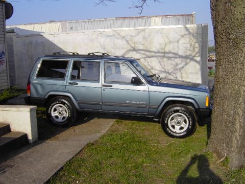 1999 Jeep Cherokee - A blue 1999 Jeep Cherokee is parked next to a tree and a building.