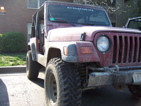 1998 Jeep TJ - A muddy red Jeep Wrangler is parked outside a brick building, showing its front grille and tires.