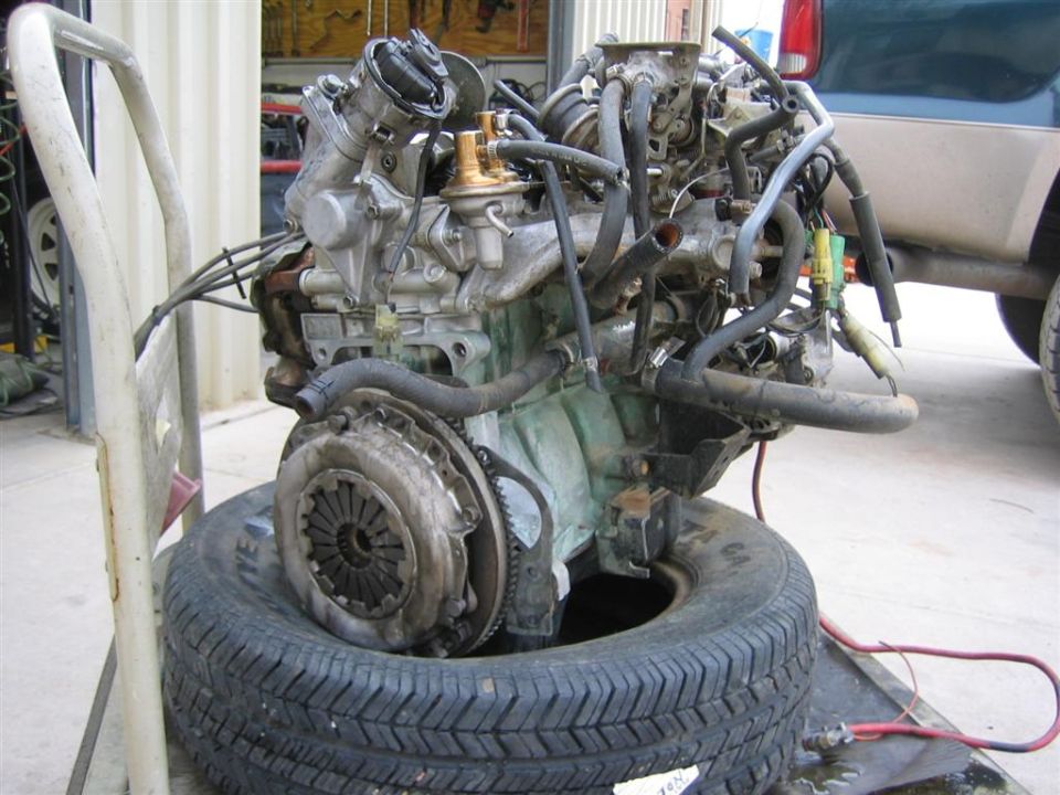 Engine and transmission assembly on a tire, with hoses and wires attached. A partial vehicle is visible in the background