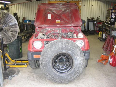 1987 Suzuki Samurai - A red SUV sits in a garage with its hood open and a large tire obscuring the front grille.