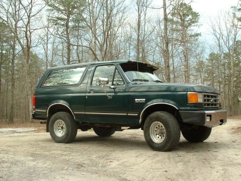 1988 Ford Bronco - This green Ford Bronco is parked outdoors on a dirt road, surrounded by trees.