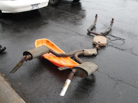 1981 Chevrolet Corvette - An exhaust system lies on a snow sled next to a white car on a wet asphalt surface.