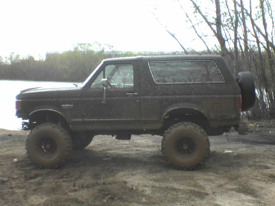 Side view of a black 1991 Ford Bronco with large tires, parked on a dirt and mud surface near water and trees