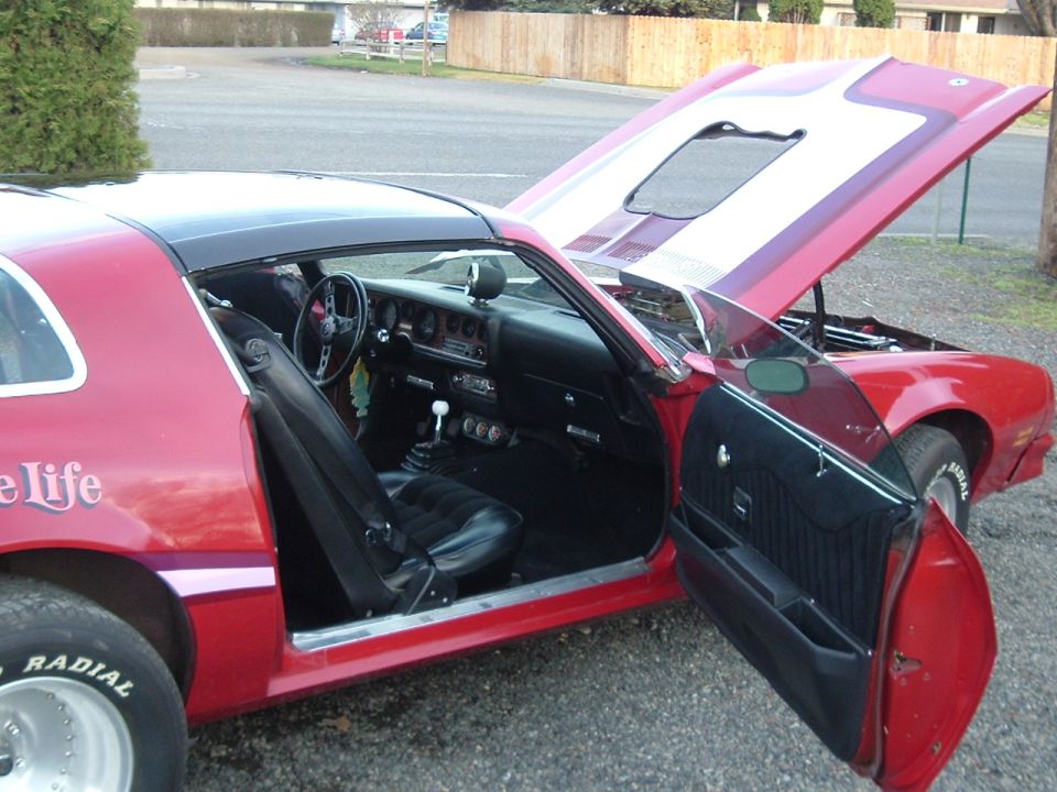 Red Firebird coupe, passenger door open, hood up, black interior with manual shifter and white hood stripe.