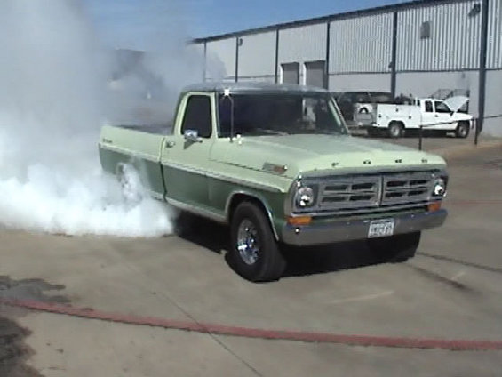 Two-tone green classic Ford pickup doing a burnout with white tire smoke on a concrete lot