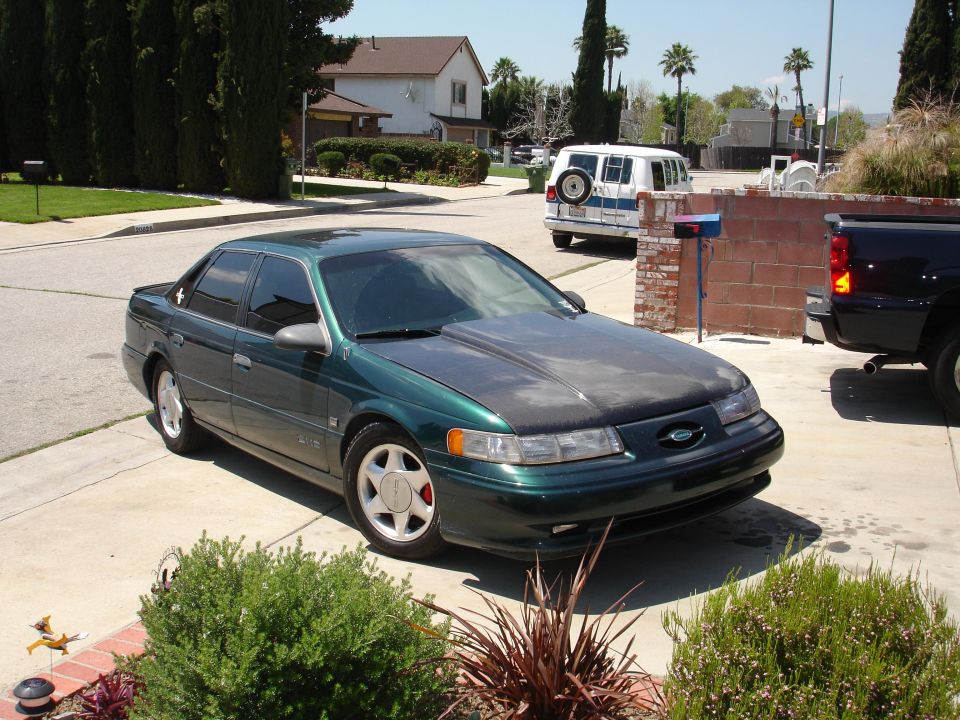 Green 1993 Ford Taurus parked in driveway, aftermarket hood, with a white van and dark pickup truck visible