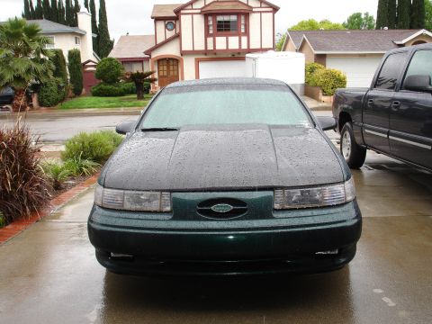1993 Ford Taurus - A green sedan with a dark hood sits on a wet driveway, with a pickup truck parked to its right.