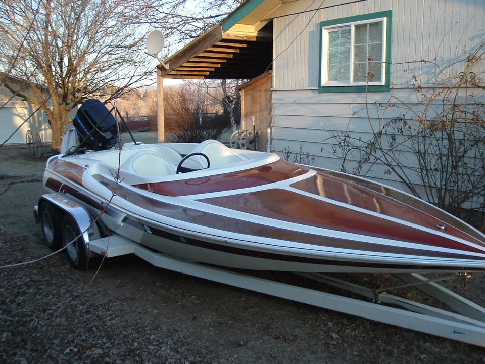 White and maroon speedboat with outboard motor on a trailer parked beside a house.