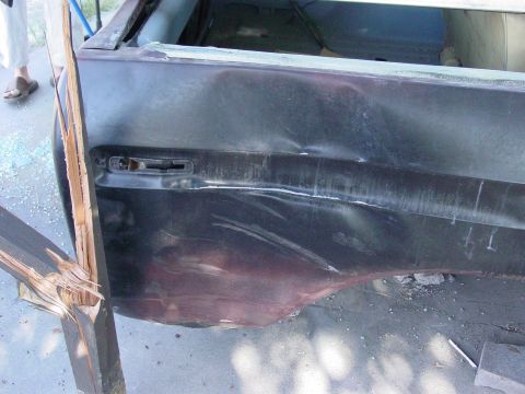 1979 Ford Bronco - The rear quarter panel of a dark vehicle exhibits extensive damage, including dents and a splintered wooden plank impacting the body.