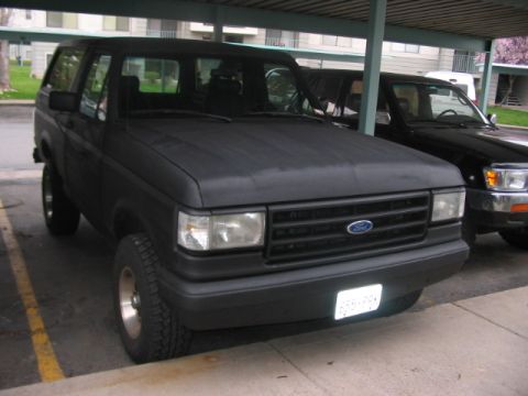 1991 Ford Bronco - A matte black Ford Bronco is parked under a covered area, with another vehicle visible to its right.