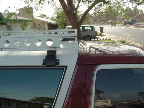 1990 Ford Bronco - Close-up of a white roof rack basket with round lightening holes mounted on the white top of a dark-maroon truck, showing a black hinged mo…
