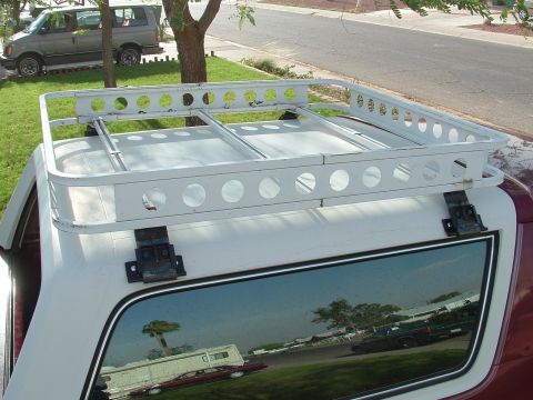1990 Ford Bronco - Overhead view of a white steel roof basket with round lightening holes bolted via black brackets to the roof of a white camper shell on a d…