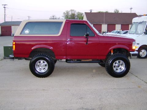 1989 Ford Bronco - Driver-side profile of a red two-door Ford Bronco with a tan removable hardtop, lifted on polished alloy wheels with tall mud-terrain tires…