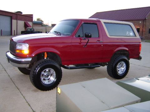 1989 Ford Bronco - Front three-quarter driver-side view of a red two-door Ford Bronco with a tan removable hardtop and its parking lamps lit, parked on concre…