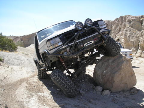 1988 Ford Bronco - This modified 1987 Ford Bronco navigates a large rock obstacle on a dusty trail, with a second vehicle visible in the distance.