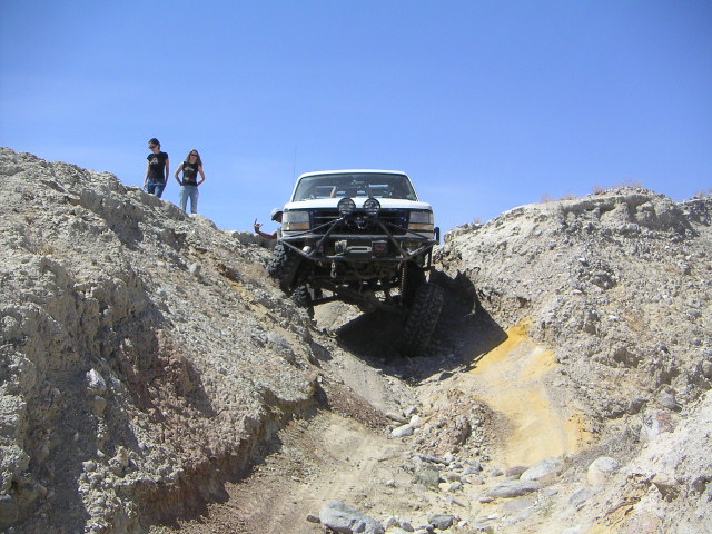 White truck with off-road bumper climbing a steep, rocky hill. Two people stand on a ridge above