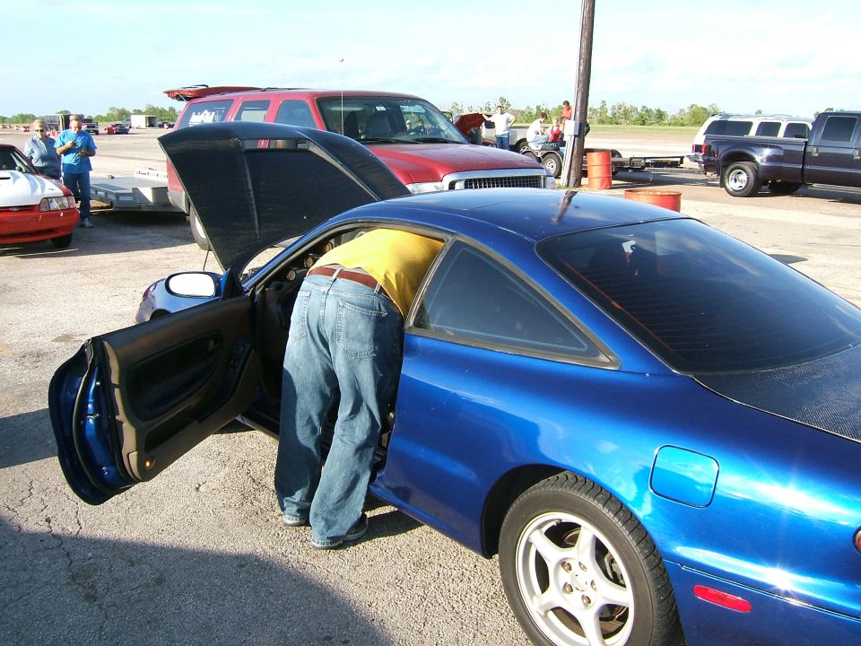Blue coupe with driver's door open, person leaning inside. Red SUV and pickup trucks in background