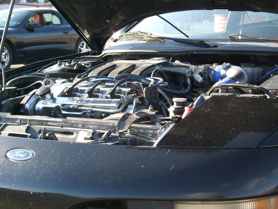 Black car engine bay with Ford logo visible. Another car is in the background