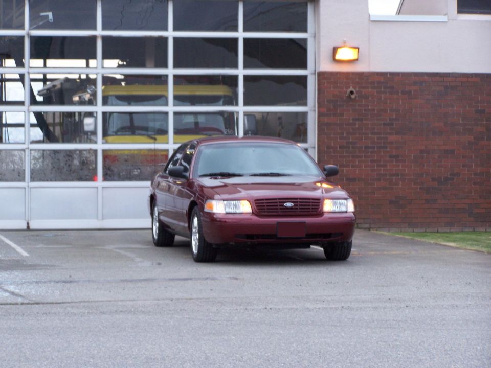 Maroon Ford sedan parked front-3/4 in front of a fire-station bay door