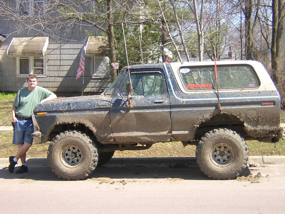 Mud-covered lifted dark blue Ford Bronco parked curbside, side view, with owner leaning on the front fender
