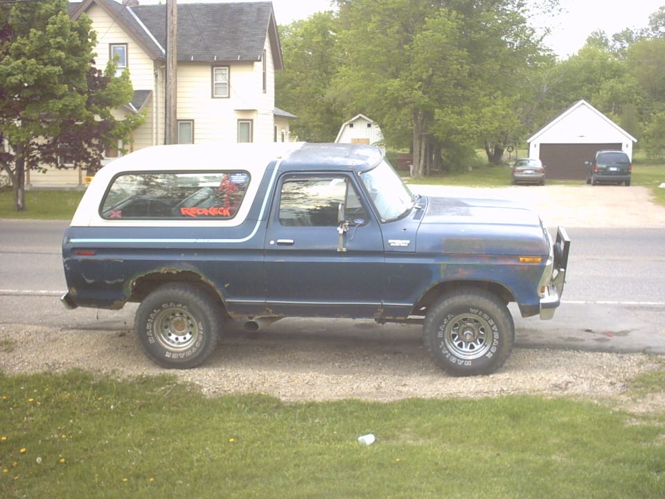 Side view of a dark blue Ford Bronco with white roof, white-letter tires, and rust, parked on a gravel roadside.