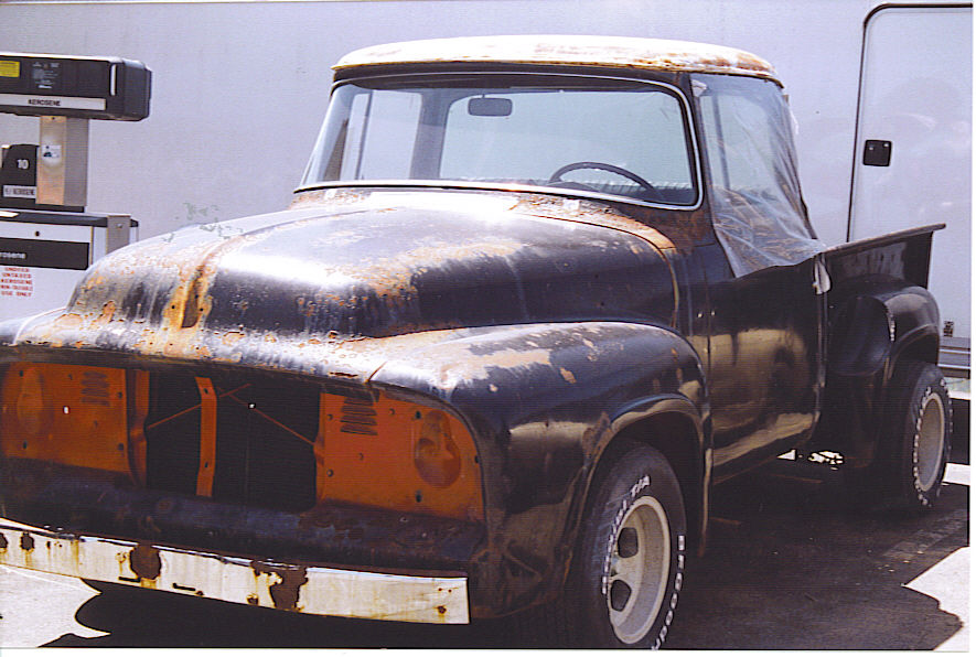 Rusty black 1956 Ford F-100 truck with a yellow roof, missing grille, and plastic covering the passenger side window