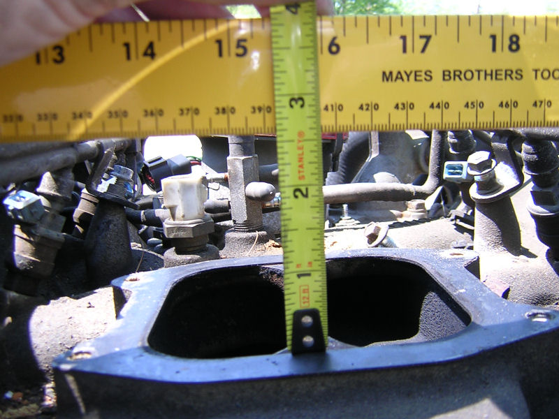 Measuring the opening of an engine intake manifold with a tape measure. The manifold has several sensors and hoses attached