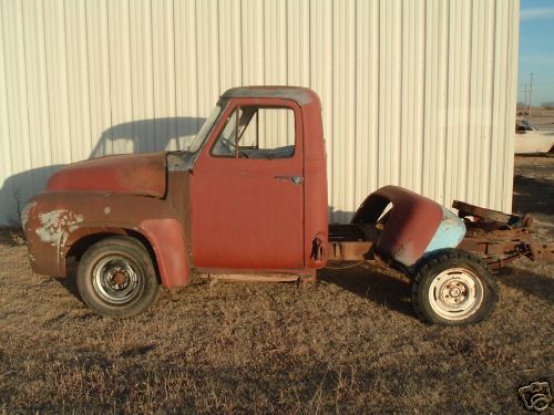 1955 Ford F-100 chassis, rusty red cab, no bed, parked outside