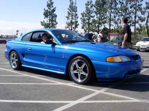 1998 Ford Crown Victoria - A blue Ford Mustang is parked in a lot with a driver inside and people standing around.