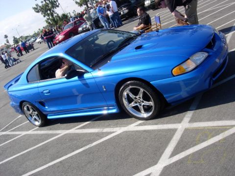 1998 Ford Crown Victoria - A bright blue Ford Mustang is parked in a lot with other cars and people around.