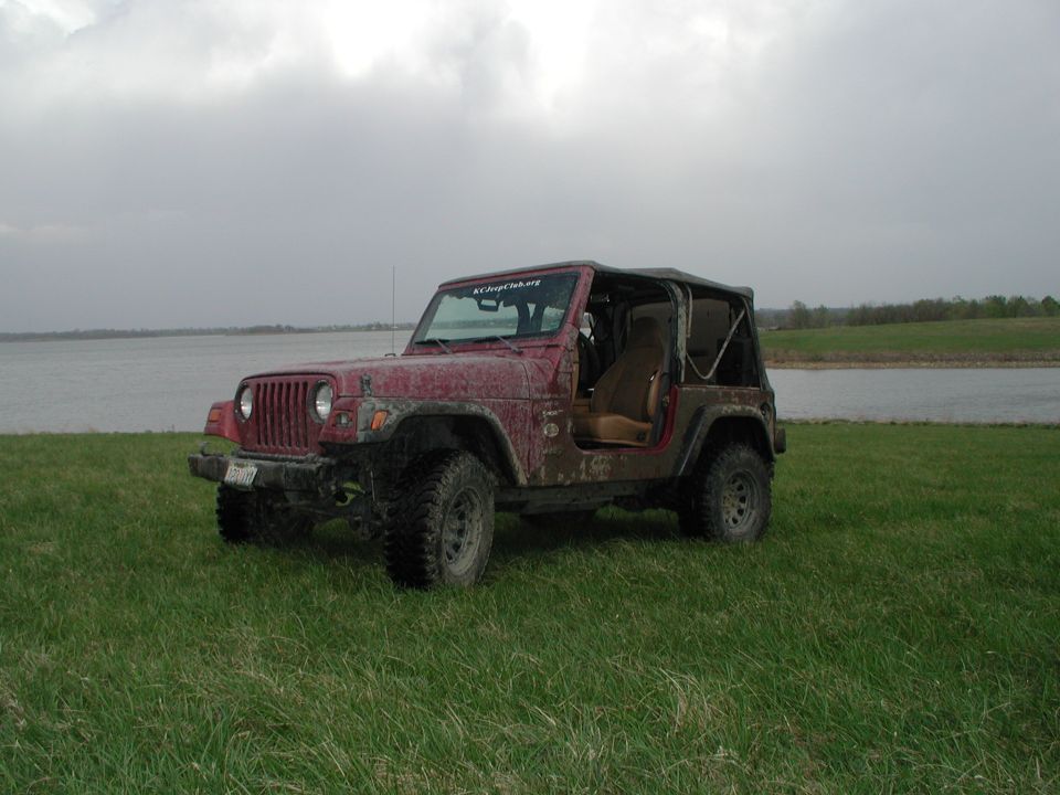 Red Jeep Wrangler parked on grass near a body of water, soft top removed